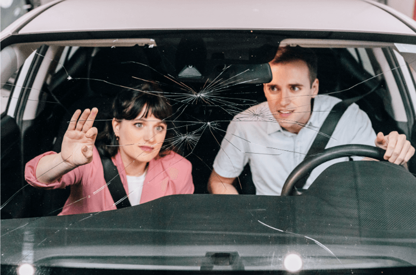 a man and woman sitting inside of a white car and staring at windshield with several cracks